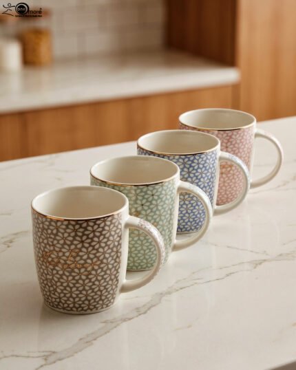 Four ceramic coffee mugs with pastel floral patterns in grey, green, blue, and pink featuring gold rims on a marble kitchen countertop.