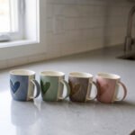 Four ceramic coffee mugs lined up on a kitchen counter in blue, green, brown, and pink with textured dot patterns and gold heart details.