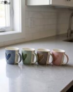 Four ceramic coffee mugs lined up on a kitchen counter in blue, green, brown, and pink with textured dot patterns and gold heart details.