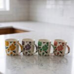 Four ceramic coffee mugs with colorful floral patterns in yellow, purple, green, and pink lined up on a marble countertop.