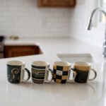 A row of four modern ceramic coffee mugs with black, cream, and brown coffee house patterns lined up on a white kitchen countertop.