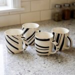 Set of 4 white ceramic coffee mugs featuring modern black stripes and subtle gold rim accents sitting on a granite kitchen countertop.