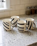 Set of 4 white ceramic coffee mugs featuring modern black stripes and subtle gold rim accents sitting on a granite kitchen countertop.