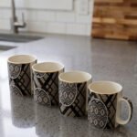 4-piece ceramic coffee mug set with black and grey geometric tribal patterns and gold rims lined up on a granite kitchen countertop