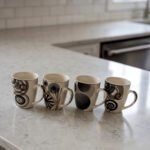 Set of 4 cream ceramic coffee mugs with distinct black geometric and swirl patterns displayed on a white marble countertop