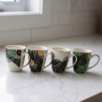 Four white ceramic coffee mugs lined up on a marble countertop with green, gold, and black abstract tribal patterns and comfortable handles.