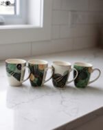 Four white ceramic coffee mugs lined up on a marble countertop with green, gold, and black abstract tribal patterns and comfortable handles.