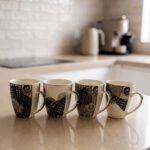 Set of 4 white ceramic coffee mugs featuring modern black geometric patterns lined up on a kitchen countertop.