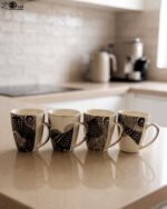Set of 4 white ceramic coffee mugs featuring modern black geometric patterns lined up on a kitchen countertop.