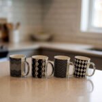 Four-piece modern black and white ceramic coffee mug set with geometric chevron and argyle patterns lined up on a kitchen counter.