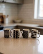 Four-piece modern black and white ceramic coffee mug set with geometric chevron and argyle patterns lined up on a kitchen counter.