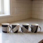 Set of 4 modern ceramic coffee mugs with distinct black, white, and grey geometric patterns displayed on a kitchen countertop.