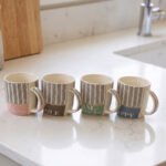 Four ceramic coffee mugs with black herringbone chevron patterns and colorful inspirational text labels lined up on a marble kitchen countertop.