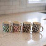 Four ceramic coffee mugs with gingham and polka dot patterns lined up on a marble countertop