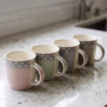 4-piece ceramic mug set in pink, green, and grey with polka dots, horizontal stripes, and gold rims on a kitchen countertop