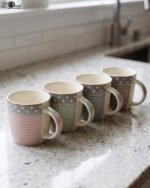 4-piece ceramic mug set in pink, green, and grey with polka dots, horizontal stripes, and gold rims on a kitchen countertop