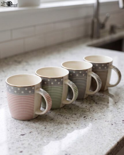 4-piece ceramic mug set in pink, green, and grey with polka dots, horizontal stripes, and gold rims on a kitchen countertop