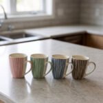 Four ceramic coffee mugs with pastel vertical stripes in pink, green, blue, and brown sitting on a marble kitchen countertop.