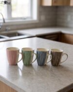 Four ceramic coffee mugs with pastel vertical stripes in pink, green, blue, and brown sitting on a marble kitchen countertop.