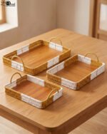 Set of three rectangular handwoven rattan serving trays with wooden bases and handles on a wooden table.