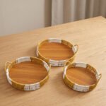 Three-piece round handwoven rattan serving tray set with white and tan woven edges and wooden bases on a light wood table.
