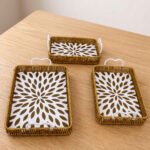 Three rectangular woven rattan serving trays with white decorative floral bottoms and side handles on a wooden table.