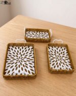 Three rectangular woven rattan serving trays with white decorative floral bottoms and side handles on a wooden table.