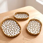 Set of 3 oval woven rattan serving trays with white handles and a modern geometric floral pattern base resting on a wooden table.