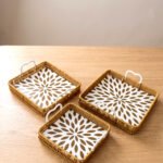 3-piece handwoven rattan serving tray set with white floral printed base and white handles on a wooden table.