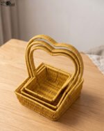 Three yellow handwoven rattan baskets nested together featuring prominent heart-shaped top handles on a wooden table.