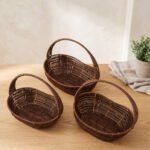 Set of three handwoven dark brown oval rattan baskets with handles resting on a light wooden table.