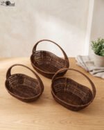 Set of three handwoven dark brown oval rattan baskets with handles resting on a light wooden table.