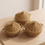 3-piece handwoven natural rattan basket set with fitted lids and side handles sitting on a light wooden table