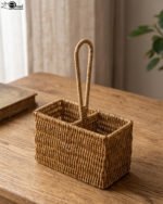 Handwoven rattan spoon holder basket with center handle and two compartments on a wooden table.