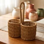 Handmade woven rattan spoon holder with two round caddy compartments and a high center handle on a wooden dining table.