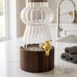 Elegant 3L ribbed glass drink dispenser sitting on a dark wooden base with a gold spigot on a kitchen counter