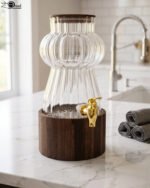 Elegant 3L ribbed glass drink dispenser sitting on a dark wooden base with a gold spigot on a kitchen counter