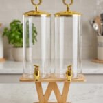 Two tall clear glass drink dispensers with gold spigots and circular gold finial lids resting on a wooden W-shaped stand.