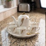 White and gold ceramic tea set with 6 cups, 1 kettle, and a rotating tray displayed on a kitchen counter.