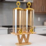 Dual 3.3L amber shaded glass beverage dispensers with gold lids and spigots on a W-shaped wooden stand sitting on a modern kitchen counter.