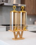 Dual 3.3L amber shaded glass beverage dispensers with gold lids and spigots on a W-shaped wooden stand sitting on a modern kitchen counter.