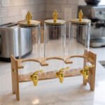 An elegant 3-tier beverage dispenser set featuring clear glass canisters with gold spigots and lids, resting on a natural bamboo stand on a white marble countertop.