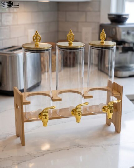 An elegant 3-tier beverage dispenser set featuring clear glass canisters with gold spigots and lids, resting on a natural bamboo stand on a white marble countertop.