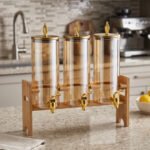 A modern kitchen counter with a three-vessel amber glass beverage dispenser set, held in a bamboo wooden stand. Each 1.8L cylinder has a gold pineapple lid and gold spigot, filled with amber liquid.
