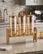 A modern kitchen counter with a three-vessel amber glass beverage dispenser set, held in a bamboo wooden stand. Each 1.8L cylinder has a gold pineapple lid and gold spigot, filled with amber liquid.