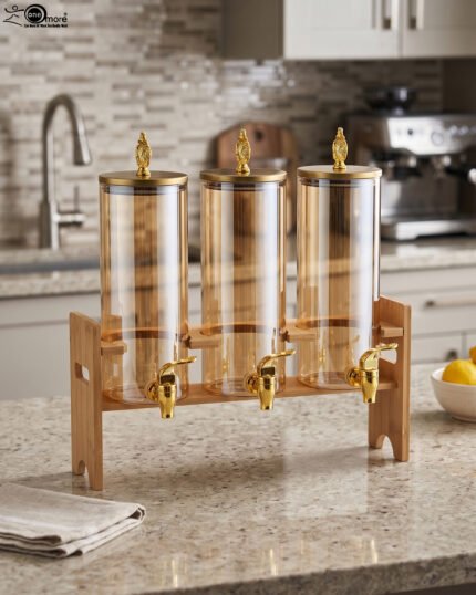 A modern kitchen counter with a three-vessel amber glass beverage dispenser set, held in a bamboo wooden stand. Each 1.8L cylinder has a gold pineapple lid and gold spigot, filled with amber liquid.