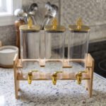 Triple clear acrylic beverage dispenser set on a wooden stand with gold spigots and decorative finial tops on a kitchen counter
