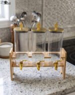 Triple clear acrylic beverage dispenser set on a wooden stand with gold spigots and decorative finial tops on a kitchen counter