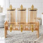 Triple 2.2L amber glass beverage dispenser set with ornate gold spigots and lids on a natural wooden stand on a marble countertop.