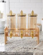 Triple 2.2L amber glass beverage dispenser set with ornate gold spigots and lids on a natural wooden stand on a marble countertop.